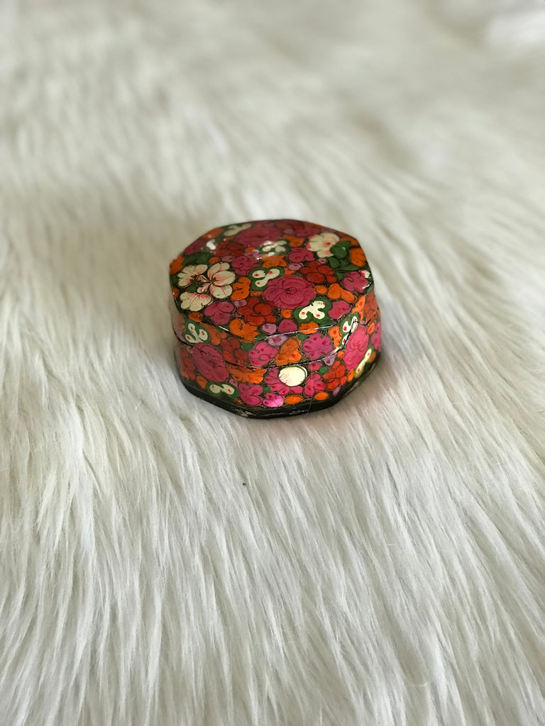 A vintage, rounded-octagonal, lidded box with hand-painted bright pink, orange, and white floral design and a black, lacquered interior. Triangle-shaped notch closure and small, faded "Made in India" sticker on bottom.