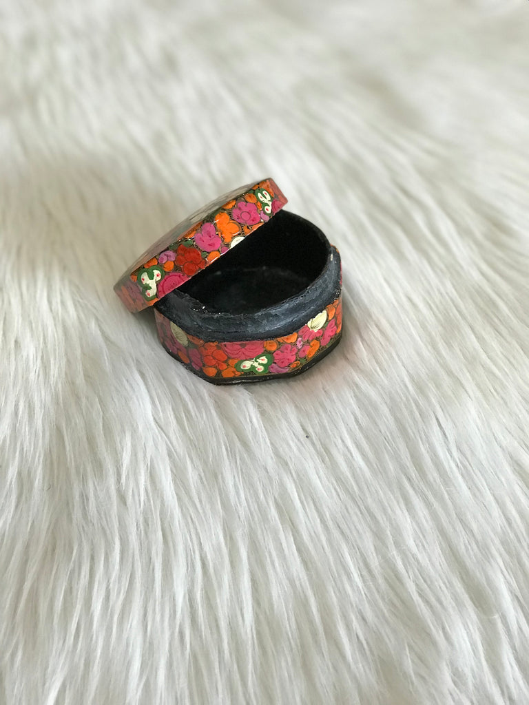 A vintage, rounded-octagonal, lidded box with hand-painted bright pink, orange, and white floral design and a black, lacquered interior. Triangle-shaped notch closure and small, faded "Made in India" sticker on bottom.